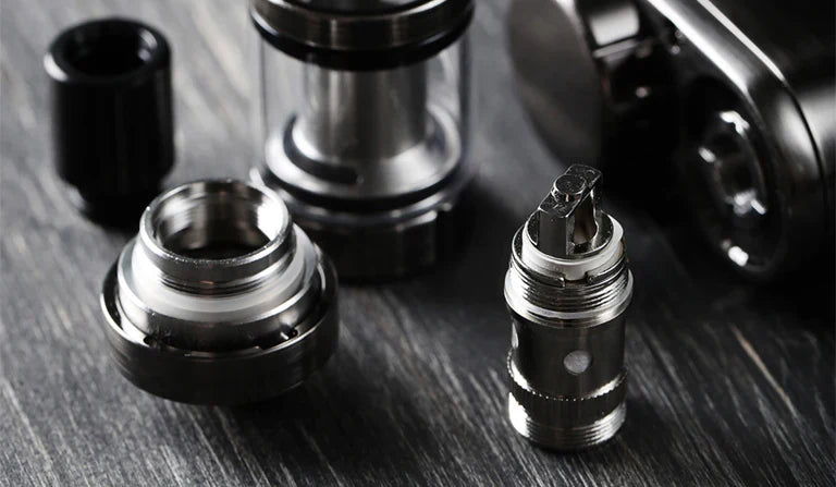 A close-up of disassembled vape tank components, including a silver atomizer coil, base, and glass tank section, resting on a dark wood surface.