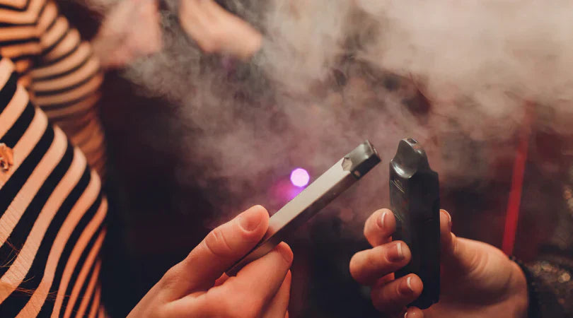 Two people holding vape devices surrounded by smoke in a dimly lit environment.