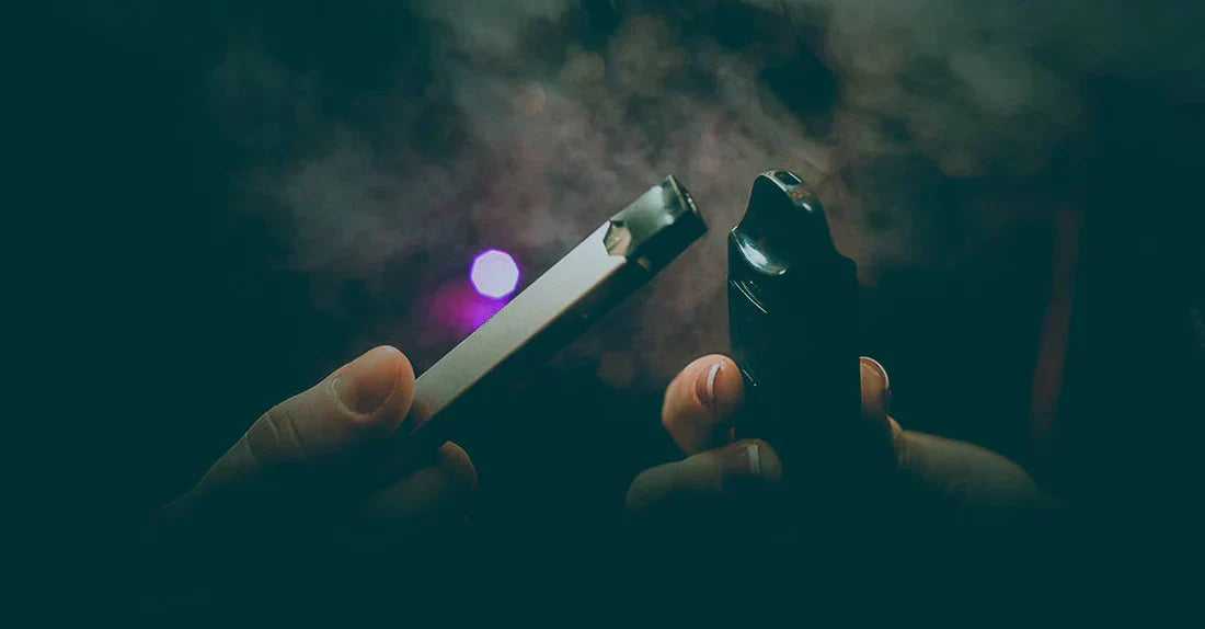 Two hands holding different vape devices are shown close together in a dark, smoky atmosphere with a purple light in the background. The scene emphasizes the act of vaping and the variety of vape devices.